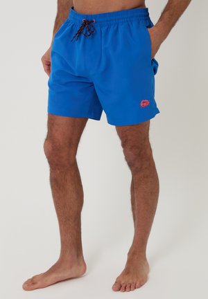 Mężczyzna w jasnoniebieskich kąpielówkach z elastycznym pasem i czarnym sznurkiem, stojący boso na jednolitym tle.