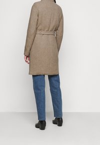 Cappotto in misto lana marrone con vita regolabile, caratterizzato da una linea sartoriale, maniche lunghe e orlo dritto, abbinato a jeans in denim blu.