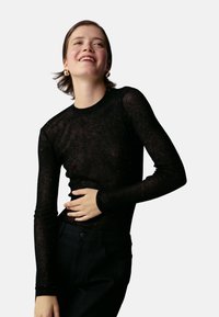 Top noir à manches longues en tissu transparent avec des paillettes subtiles. Silhouette ajustée, col rond et poignets côtelés. Assorti à un pantalon foncé.