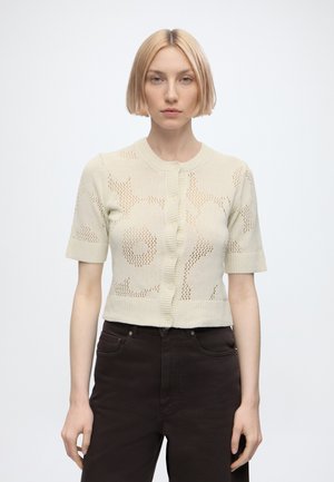 Femme aux cheveux blonds courts portant un cardigan beige à manches courtes tricoté et un pantalon taille haute foncé sur fond blanc uni.