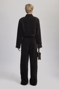 Hnědá sametová zkrácená bunda a kalhoty, které mají texturovanou látku, uvolněný střih a malou černou kabelku drženou v ruce.