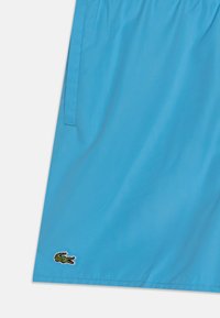 Világoskék fürdőnadrág sima anyagból, oldalsó zsebekkel és egy kicsi, zöld hímzett krokodil logóval az alsó szegély közelében.