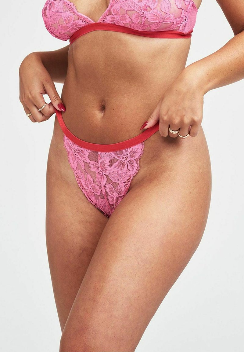 Rosa Spitzen-Bralette und String-Set mit floralen Mustern, transparentem Stoff und rotem Besatz. Der String hat ein hochgeschnittenes Design und minimalen Sitz.