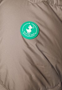 Rund grøn lap med en hvid and og teksten "RED DIN AND" på brunt stof, sandsynligvis en jakke eller frakke.