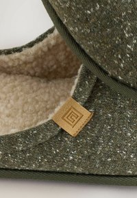Grønne uldslippers med en struktureret overflade, blød cremefarvet foring og et brunt lædermærke med et geometrisk logo.