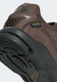 Brun atletisk sko med en strukturerad ovandel med Cordura-märkning. Såljet är svart med en böjd design och mönster för grepp.