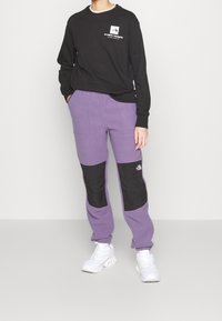 Svart långärmad tröja med logotyp, kombinerad med lila sweatpants med svarta paneler och två sidofickor. Vita träningsskor.