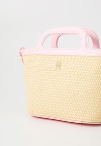 Borsa in paglia con un corpo giallo testurizzato e manici in pelle rosa. Presenta un accentuato logo dorato sul fronte. Design compatto e strutturato.