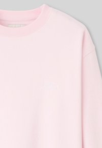 Sweat-shirt rose clair avec un col rond, des manches longues et une délicate étiquette brodée "hors série n°1" sur le devant. Texture de tissu douce.