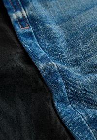 Denim tkanina v modri barvi z bledenjem in kontrastnim črnim materialom, ki prikazuje gladke teksture in vidne šive ob stikih.