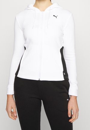 Personne portant un sweat à capuche blanc zippé avec des panneaux latéraux noirs et un pantalon noir, tous deux avec des logos Puma.