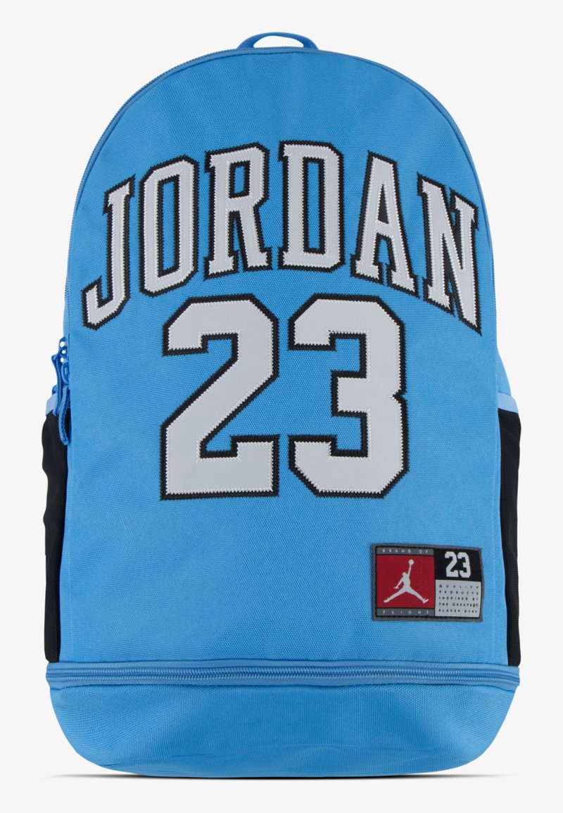 Mochila azul clara com a inscrição "JORDAN 23" em letras negritas, pretas e brancas. Inclui um bolso lateral preto e um patch de logotipo vermelho. Material durável.