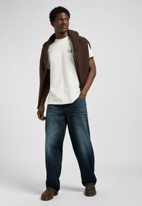 Model wearing a cream t-shirt with "LE" logo, oversized blue denim jeans, and a brown knit sweater draped over shoulders. Brown boots.