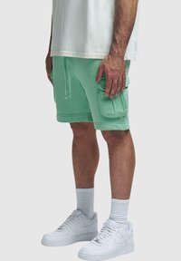 Mintgrüne Shorts mit weichem Material, ausgestattet mit seitlichen Taschen und einem Kordelzug-Bund, kombiniert mit weißen Sneakers und Socken.