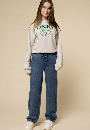 Grijze sweatshirt met de tekst "LUCKY" en een klavertje ontwerp, gecombineerd met blauwe baggy jeans. Witte sneakers maken de casual outfit compleet.