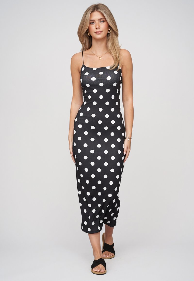 Femme aux longs cheveux blonds portant une robe midi noire à pois blancs et des sandales noires, debout devant un fond clair uni.