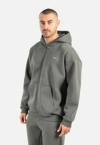Homme portant un sweat à capuche gris zippé avec poches avant et un pantalon de jogging assorti, se tenant devant un fond clair.
