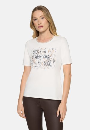 Vrouw met blond haar, gekleed in een wit kortärmelig shirt met een geometrisch patroon en de tekst "KEEP GOING", gecombineerd met donkerbruine broek.