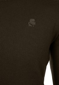 Hnědý žebrovaný pletený svetr s jemně vyraženým logem. Má kulatý výstřih a přiléhavé rukávy, což vytváří strukturovaný vzhled.