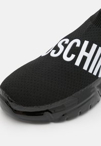 Baskets slip-on en maille noire avec semelle brillante présentant de grandes lettres blanches en relief formant partiellement un nom de marque sur le dessus.