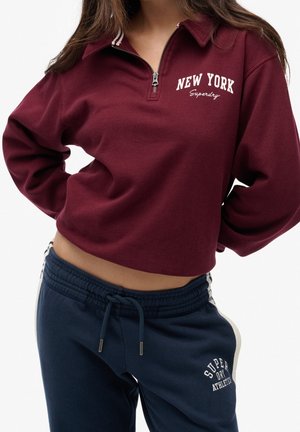 Femme portant un sweat-shirt bordeaux à fermeture éclair quart avec le texte "New York" et un pantalon de survêtement bleu marine avec cordon de serrage et logo sur la cuisse gauche.