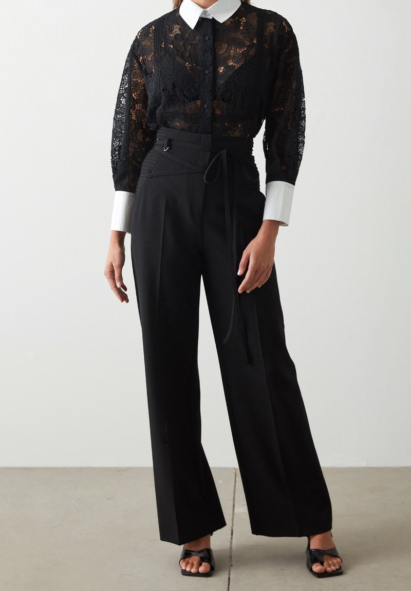 Czarna bluzka z koronką i białym kołnierzykiem, zestawiona z czarnymi spodniami o wysokim stanie z paskiem. Gładkie tekstury, dopasowany krój.