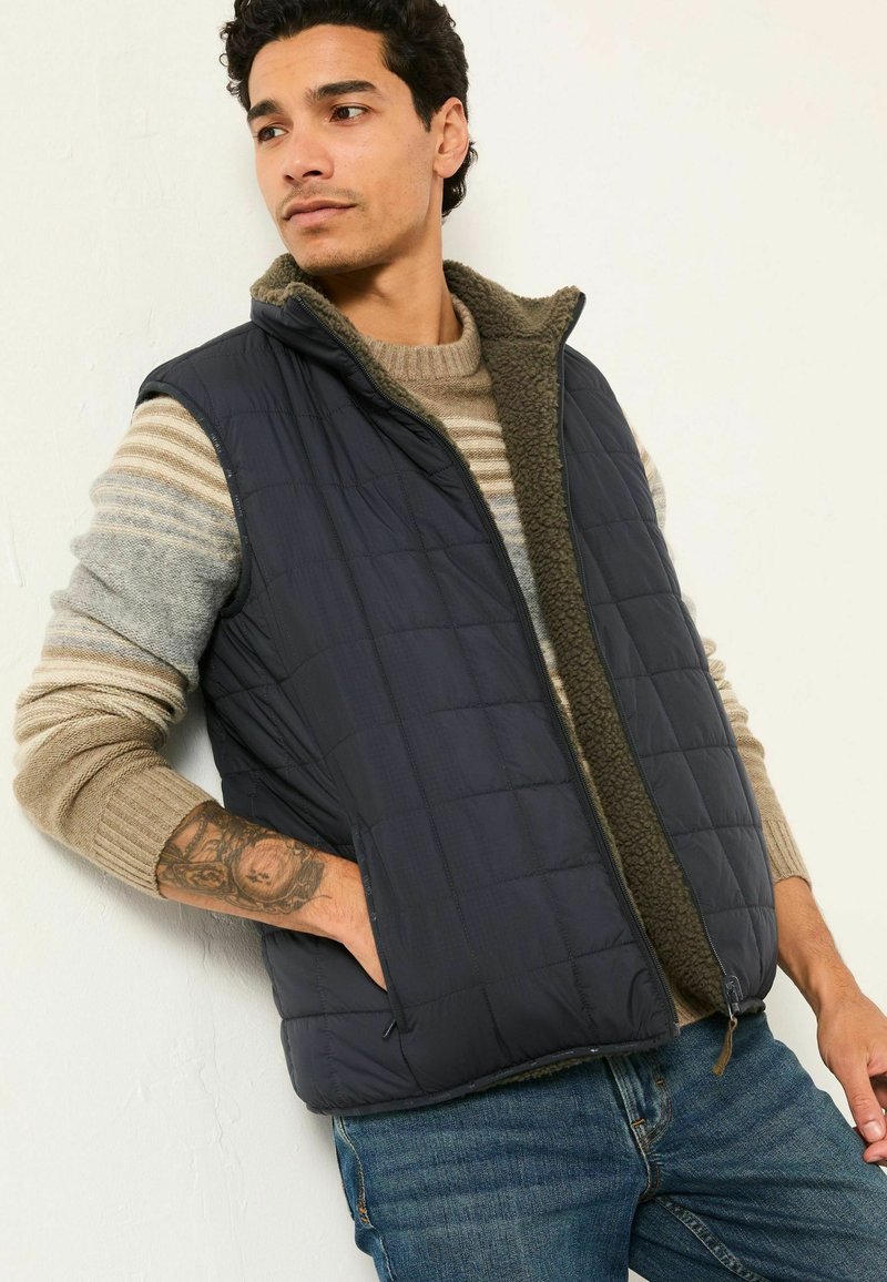 Mann trägt gestreiften beigen Pullover, dunkelblaue gesteppte Weste mit Fleece-Futter und blaue Jeans, posiert mit der Hand in der Westentasche.