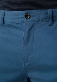 Kék pamut nadrág gombos zárással, finom textúrával és egyenes szabással, oldalsó zsebekkel és a tervezésbe integrált övhurkokkal.