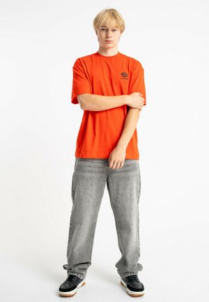 Młoda osoba o blond włosach, ubrana w pomarańczową koszulkę z krótkim rękawem, szare, luźne dżinsy oraz czarno-białe sneakersy, stoi z jedną ręką skrzyżowaną.