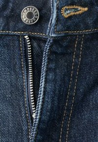 Primer plano de pantalones vaqueros de mezclilla azul con botón metálico visible, cremallera plateada parcialmente abierta y detalles de costura naranja.