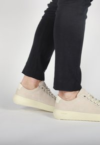 Beige suede sneakers with white rubber soles, featuring round laces and minimalistic design. Paired with cropped black pants.
