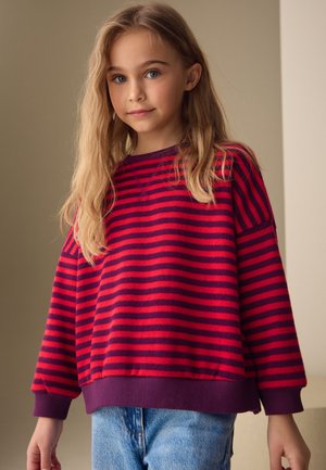 Jong meisje met lang blond haar, een rood-paars gestreepte trui en blauwe spijkerbroek, staand voor een effen beige achtergrond.
