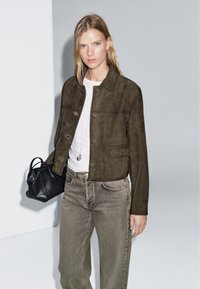 Giacca in suedè marrone con colletto, bottoni frontali e due tasche, abbinata a una t-shirt in stoffa leggera e jeans grigi, mentre tiene una borsa nera.