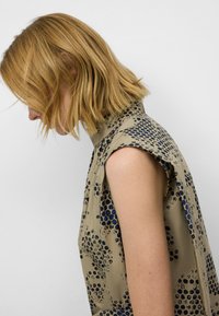 En kvinna med axellångt blont hår tittar ner, iklädd en ärmlös, beige topp med blå och svarta mönster mot en vit bakgrund.