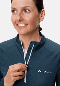 Pull-over mi-zip en teal foncé avec un tissu texturé, fermeture éclair blanche et logo. Le modèle ajuste la fermeture éclair, mettant en valeur les détails du design.