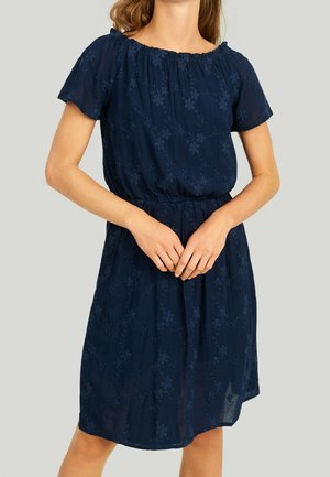 Femme portant une robe bleu marine à manches courtes et longueur genou, avec un motif floral et une taille élastique, debout devant un fond uni.