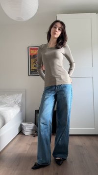 Ung kvinde står i et soveværelse iført en beige, ribstrikket, langærmet top, blå viddebenede jeans og sorte spidse sko.