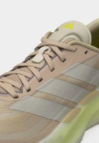 Μπεζ και γκρι αθλητικό παπούτσι Adidas με πλέγμα ύφασμα, παχιά κίτρινο-πράσινη σόλα, τρεις ρίγες στο πλάι και δεμένες κορδόνια με κλείδωμα κορδονιών.