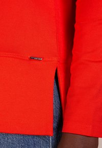 Czerwona koszula z długim rękawem wykonana z gładkiej bawełny, z bocznym rozcięciem i małą metką marki przyszytą przy dole.