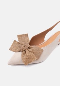 Μπεζ μπαλαρίνα με λουρί στο πίσω μέρος, μυτερή μύτη και μεγάλο υφασμάτινο χρυσό φιόγκο μπροστά, με χαμηλό τακούνι και λείο εσωτερικό επένδυση.