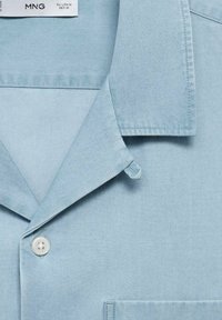 Camisa de botones color azul claro con cuello tipo solapa, un bolsillo de parche y una textura de tela suave. Incluye una etiqueta colgante.