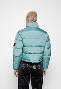 Lys teal pufferjakke med høy krage, kviltet design og elastiske mansjetter, kombinert med svarte fløyelsbukser med rhinestones.