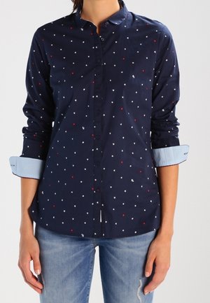 Femme portant une chemise bleu marine à boutons avec des motifs d'étoiles rouges et blanches, manches retroussées, associée à un jean bleu clair déchiré.