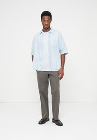 Φωτεινό γαλάζιο ριγέ κοντομάνικο πουκάμισο πάνω από ένα γαλάζιο μπλουζάκι, σε συνδυασμό με γκρι παντελόνια και σκούρα παπούτσια, στέκεται μπροστά σε ένα λευκό φόντο.