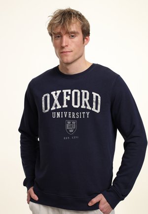 Joven con cabello claro que lleva una sudadera azul marino de la Universidad de Oxford, de pie frente a un fondo claro y liso.