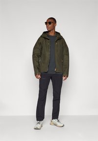 Olivgrüne, kapuzenjacke mit Frontreißverschluss und Druckknöpfen, graues T-Shirt darunter, dunkle Hose und hellfarbene Sportschuhe.