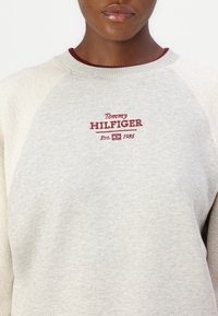 Sweatshirt gris clair avec un col rond à côtes, des accents bordeaux contrastants et un logo brodé "Tommy Hilfiger" avec "Est. 1985."