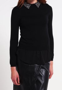 Maglione nero con colletto di perline, tessuto testurizzato, slim fit, sovrapposto a una maglietta nera con orlo a volant e bottoni. Gonna di pelle.