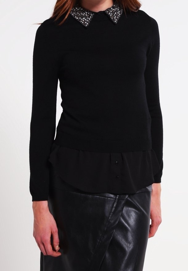 Maglione nero con colletto di perline, tessuto testurizzato, slim fit, sovrapposto a una maglietta nera con orlo a volant e bottoni. Gonna di pelle.