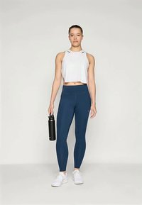 Bijela uska majica bez rukava i tamnoplave tajice viskog struka. Model drži crnu bocu za vodu, nosi bijele sportske cipele. Glatka tekstura, prilagođeni dizajn.